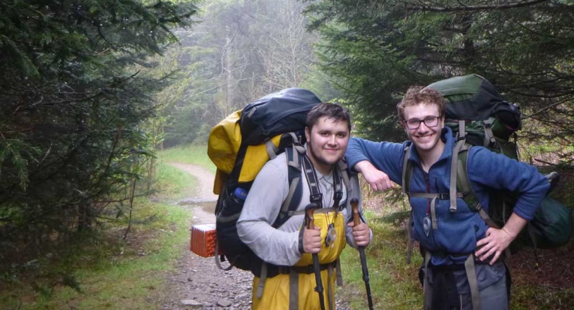 Two people wearing backpacks pose for the photo on a wooded trail. 